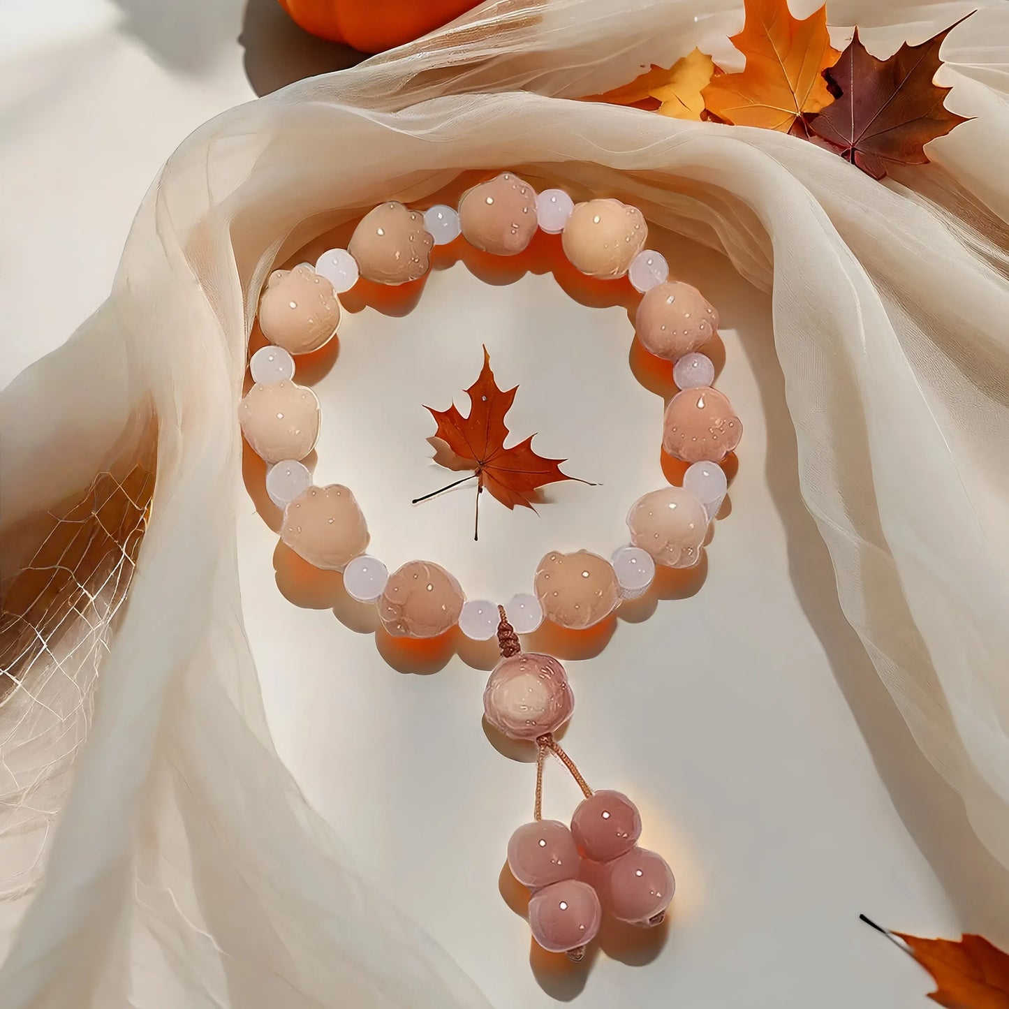 The 24 Solar Terms Series TAOÉ Pawfect Bodhi Bracelet: Carved Cat Paw Pendant - Autumnal Equinox features pink and white beads with a cat paw charm, styled on sheer fabric amid autumn leaves for a cozy, seasonal vibe.