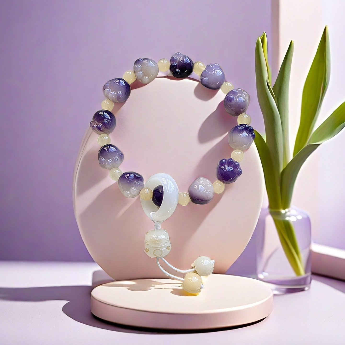 The TAOÉ Pawfect Bodhi Bracelet: Carved Cat Paw Pendant - Frosts Descent from The 24 Solar Terms Series, featuring alternating purple and white carved beads, is displayed on a pastel stand beside a leafy plant in soft natural light.