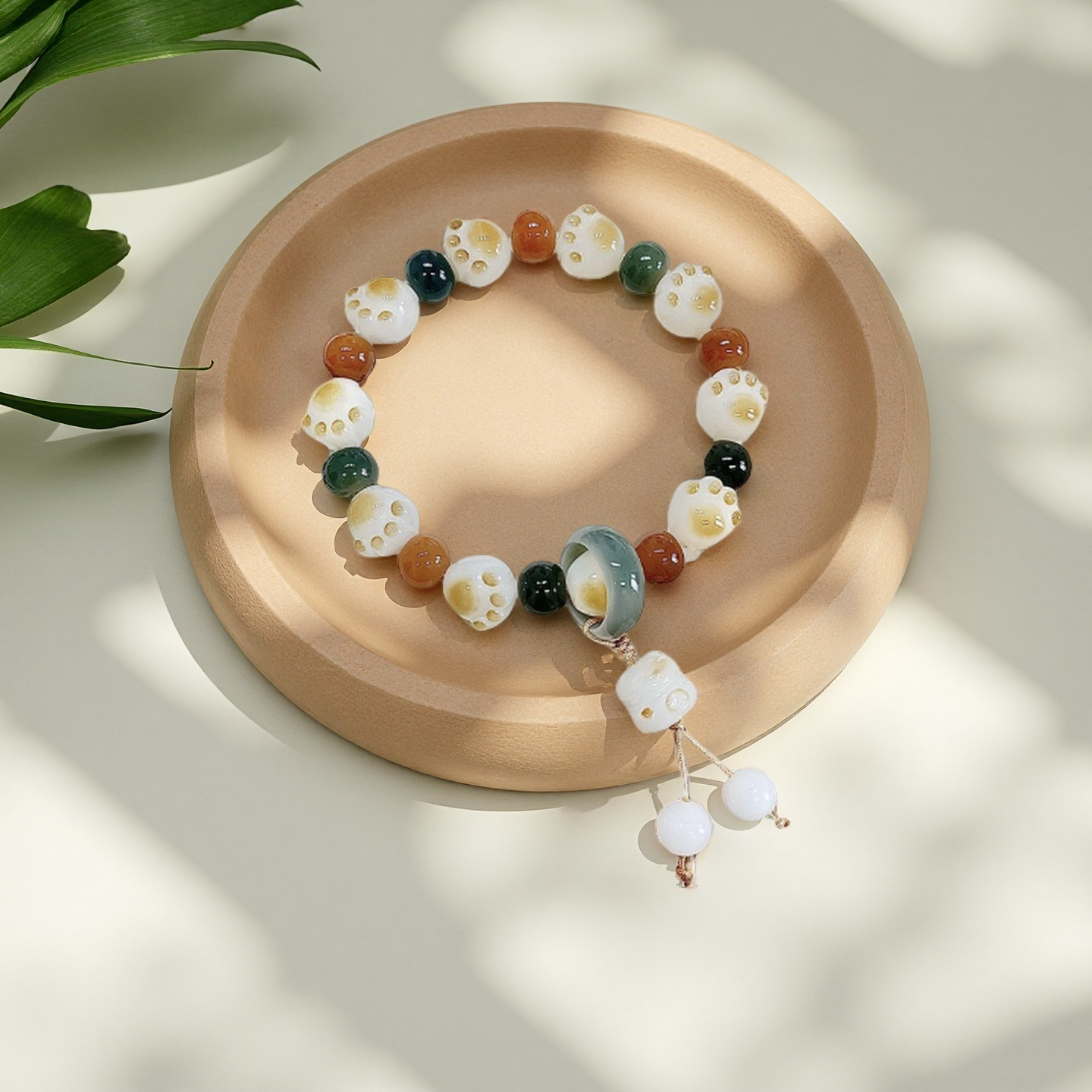 The TAOÉ Pawfect Bodhi - Start of Summer Lime Green Bracelet from The 24 Solar Terms Series features colorful round beads, white skull and cat paw beads, plus a dangling charm, displayed on a beige plate with green leaves casting shadows nearby.