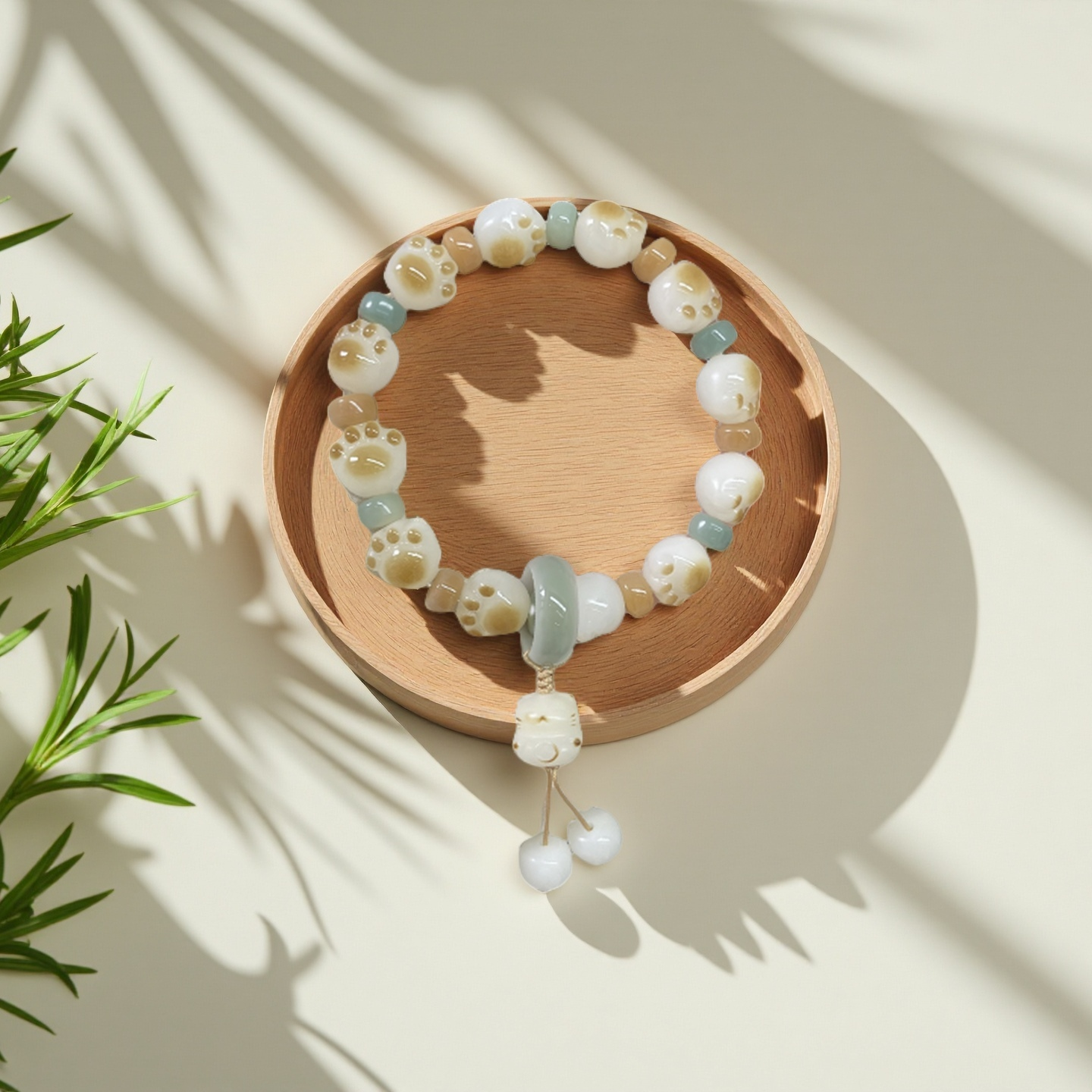 The TAOÉ Pawfect Bodhi - Winter Solstice Whisker Bracelet from The 24 Solar Terms Series, featuring white and light blue beads, sits on a round wooden dish with leafy plant shadows cast in soft natural light.