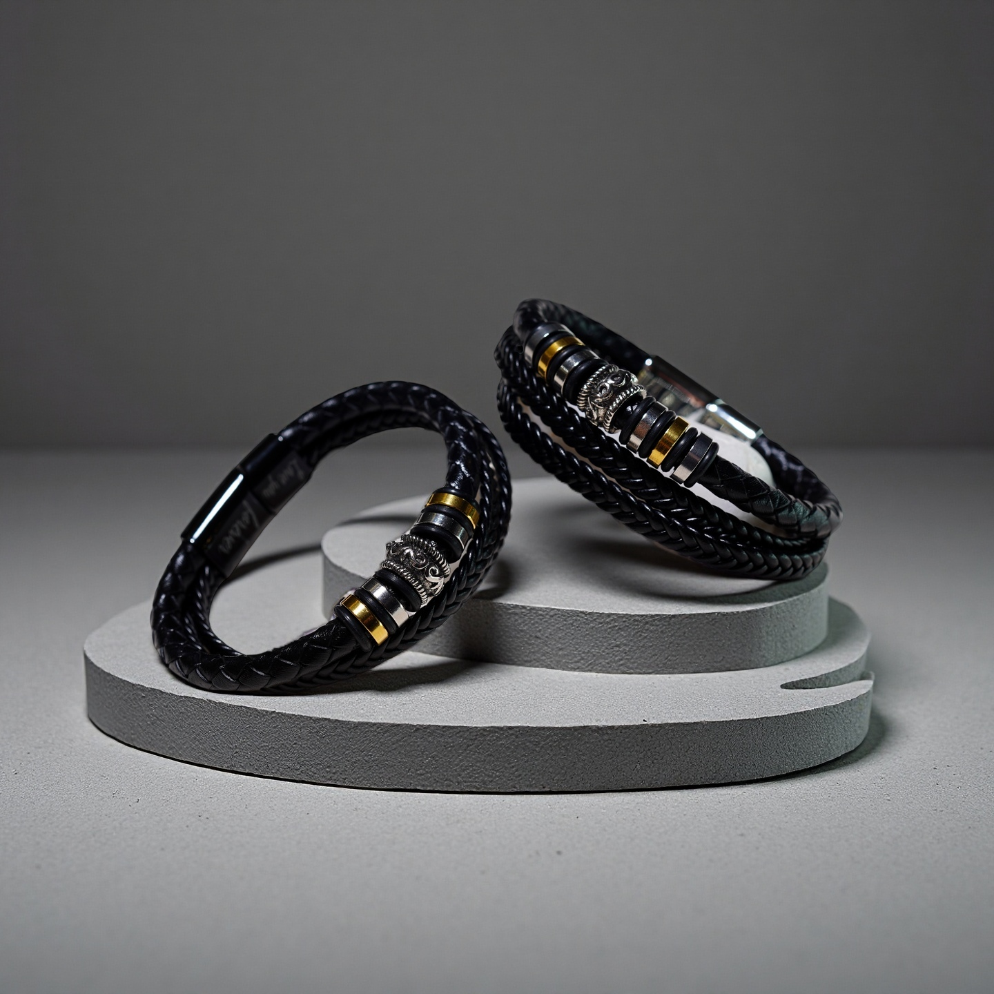 Two TAOÉ Mens Tribal Bracelets from the Five Elements Series, featuring black braided leather and stainless steel accents, are displayed on round gray stands against a neutral background.