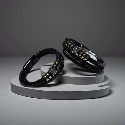Two TAOÉ Mens Tribal Bracelets from the Five Elements Series, featuring black braided leather and stainless steel accents, are displayed on round gray stands against a neutral background.