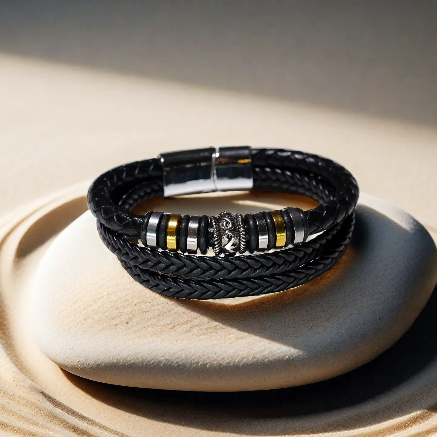 The TAOÉ Mens Tribal Bracelet from the Five Elements Series features black braided genuine leather with silver, gold, and black stainless steel accents—displayed coiled on beige stone under soft light, inspired by earth elements.