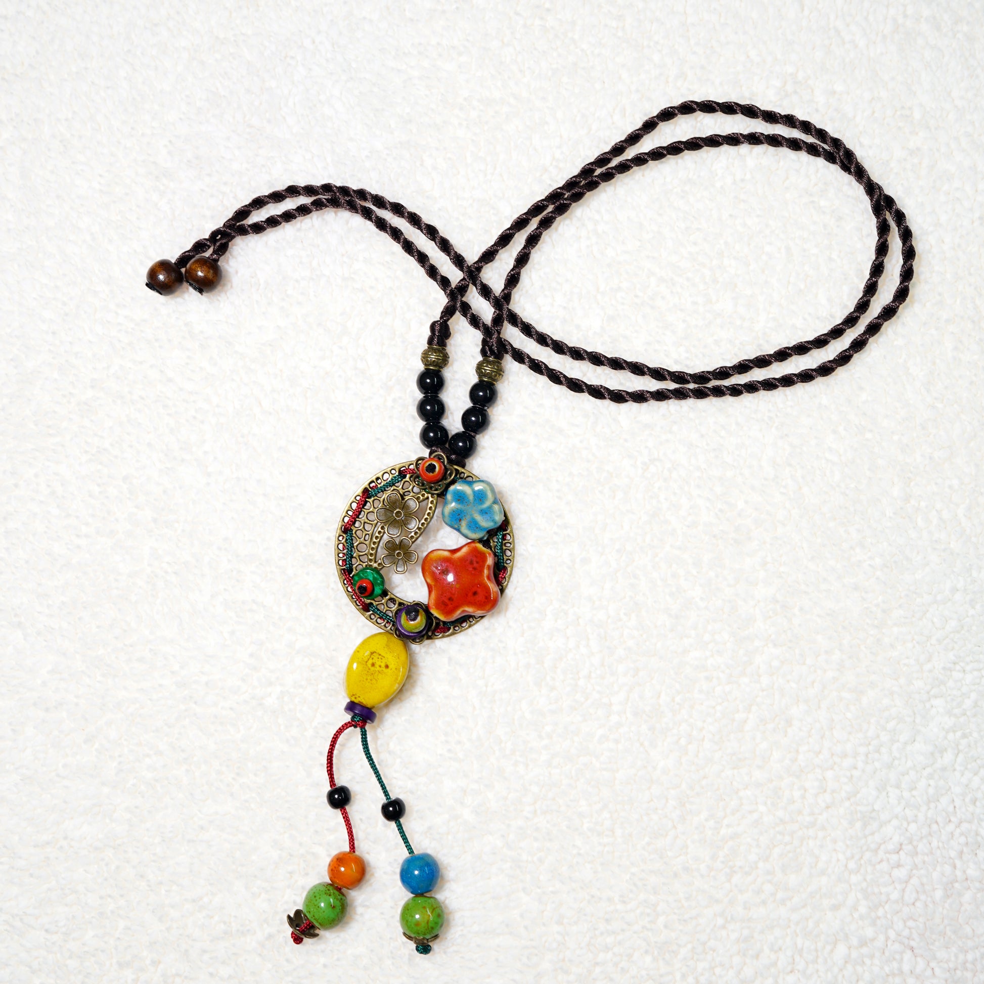 The TAOÉ Handmade Colorful Ceramic Necklace by My Store features a circular, ornate charm with red, yellow, blue, and green ceramic beads on a twisted dark brown cord, beautifully displayed on a white textured background.