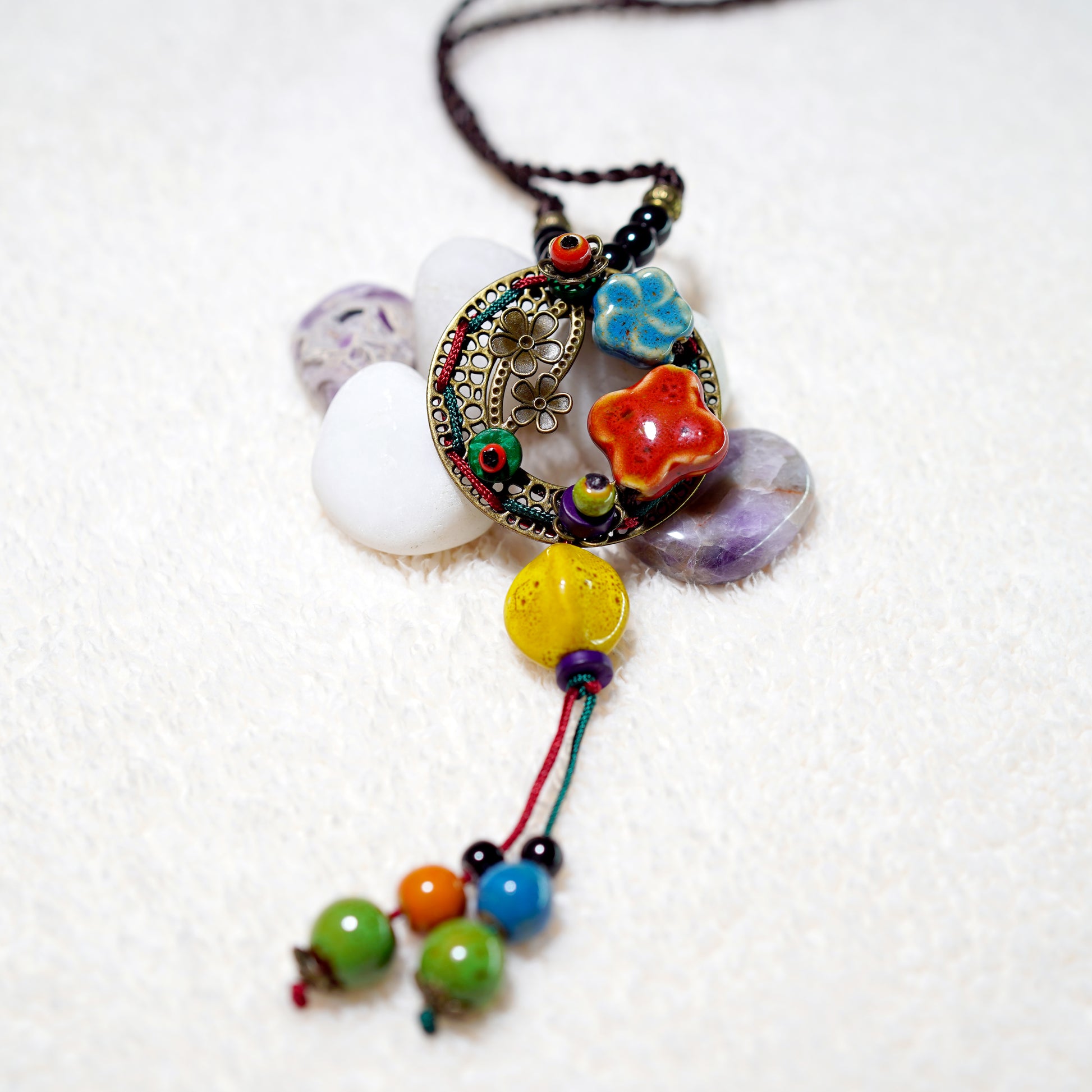 The TAOÉ Handmade Colorful Ceramic Necklace by My Store showcases artisan design with a beaded pendant, intricate metalwork, ceramic flower beads, and multicolored round beads, beautifully displayed on white stones and an off-white textured surface.
