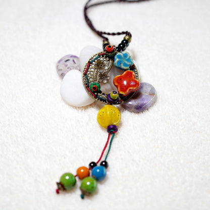 The TAOÉ Handmade Colorful Ceramic Necklace by My Store showcases artisan design with a beaded pendant, intricate metalwork, ceramic flower beads, and multicolored round beads, beautifully displayed on white stones and an off-white textured surface.